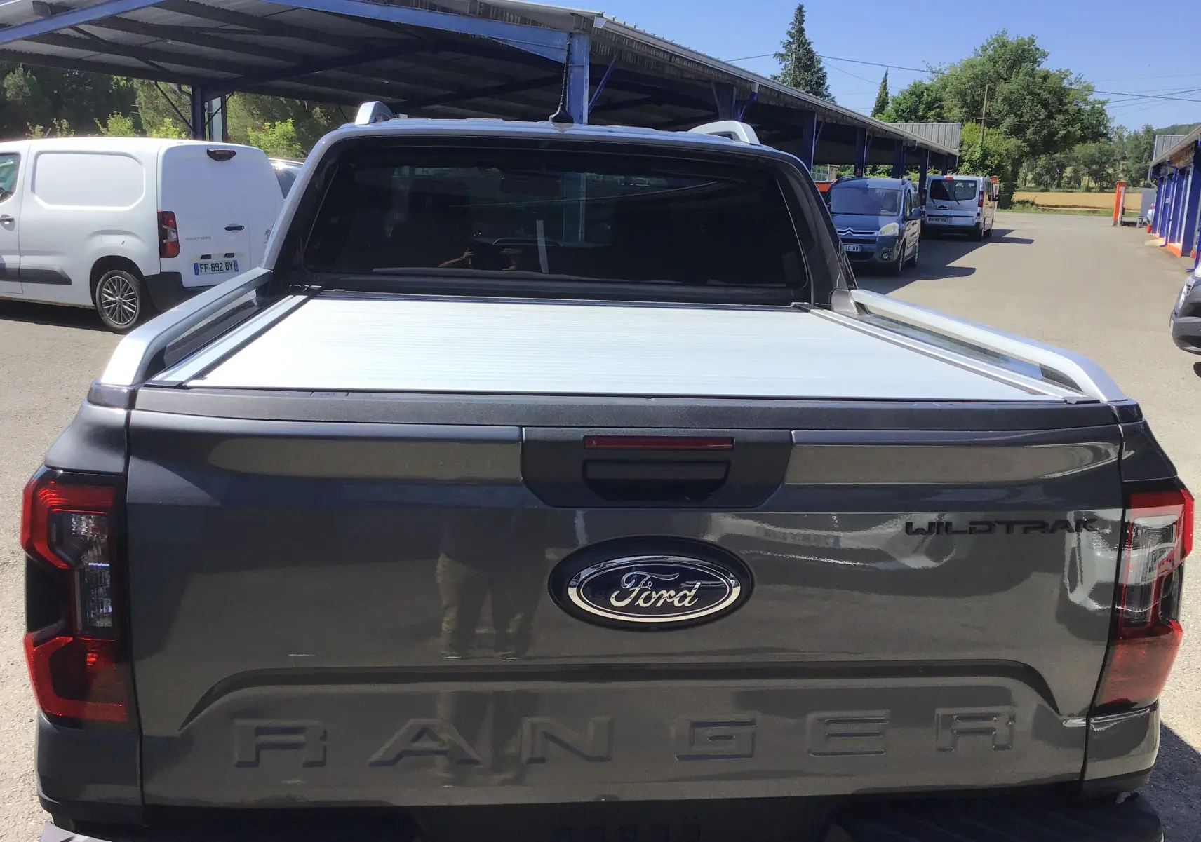 Vue arrière d'un Ford Ranger Double Cabine gris carbone avec rideau de plateau électrique et logo Wildtrak visible.