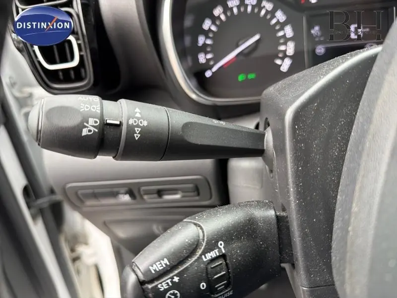 Gros plan sur les commandes de phares et régulateur de vitesse dans l'habitacle gris d'une Citroën C3 Aircross blanche.