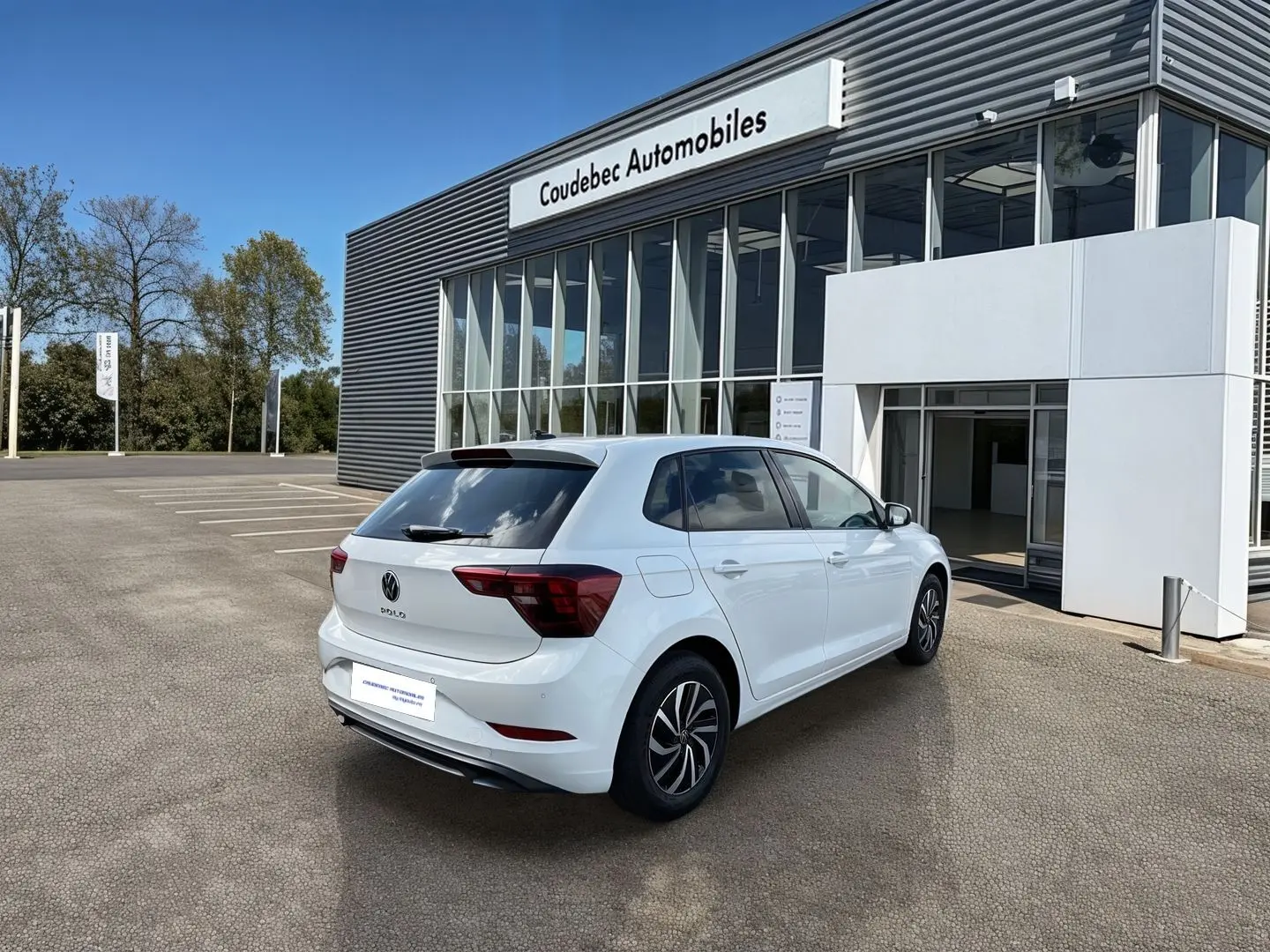 Vue 3/4 arrière droite d’une Volkswagen Polo blanche stationnée devant un bâtiment vitré de concession.