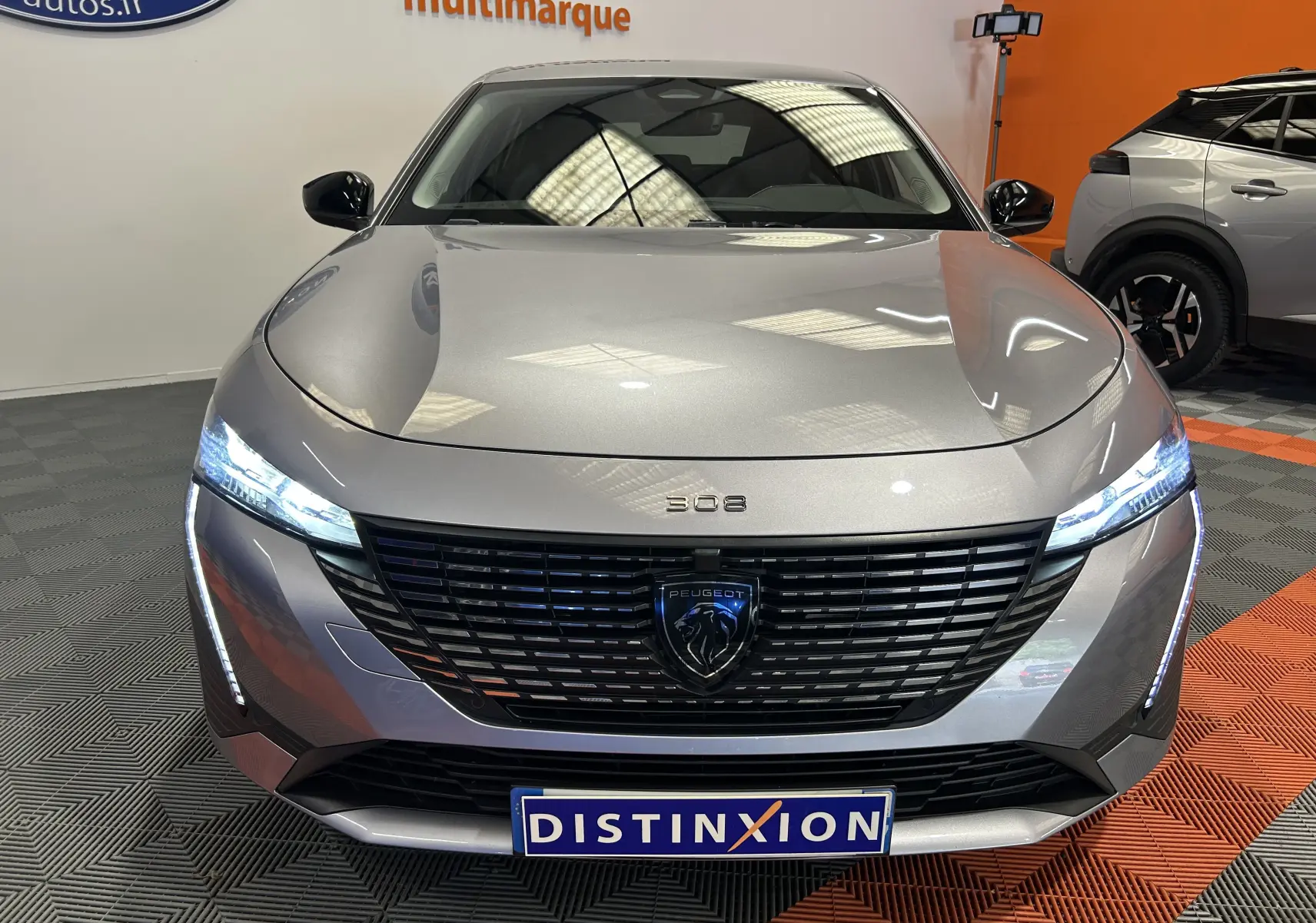 Vue frontale d'une Peugeot 308 gris métallisé avec phares LED allumés et calandre noire distinctive en showroom.