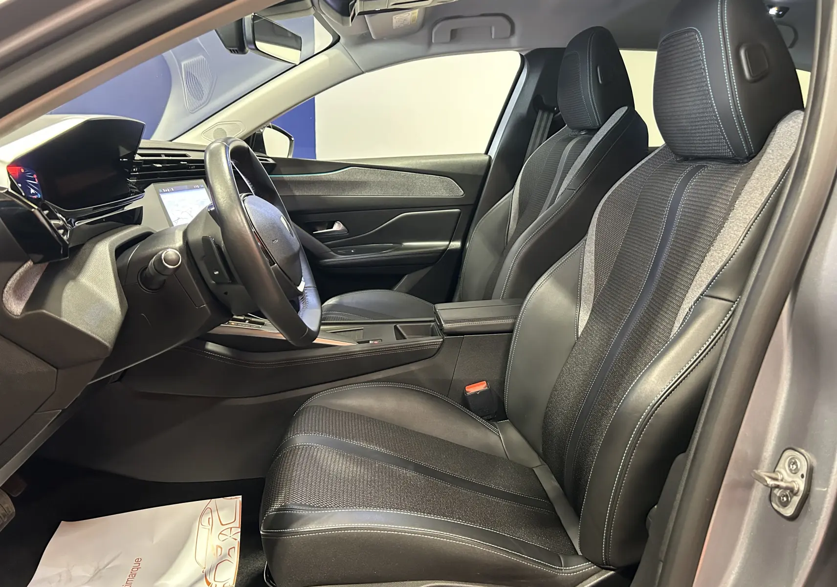 Vue intérieure côté conducteur de la Peugeot 308 gris, mettant en valeur les sièges en cuir et tissu avec surpiqûres bleues.
