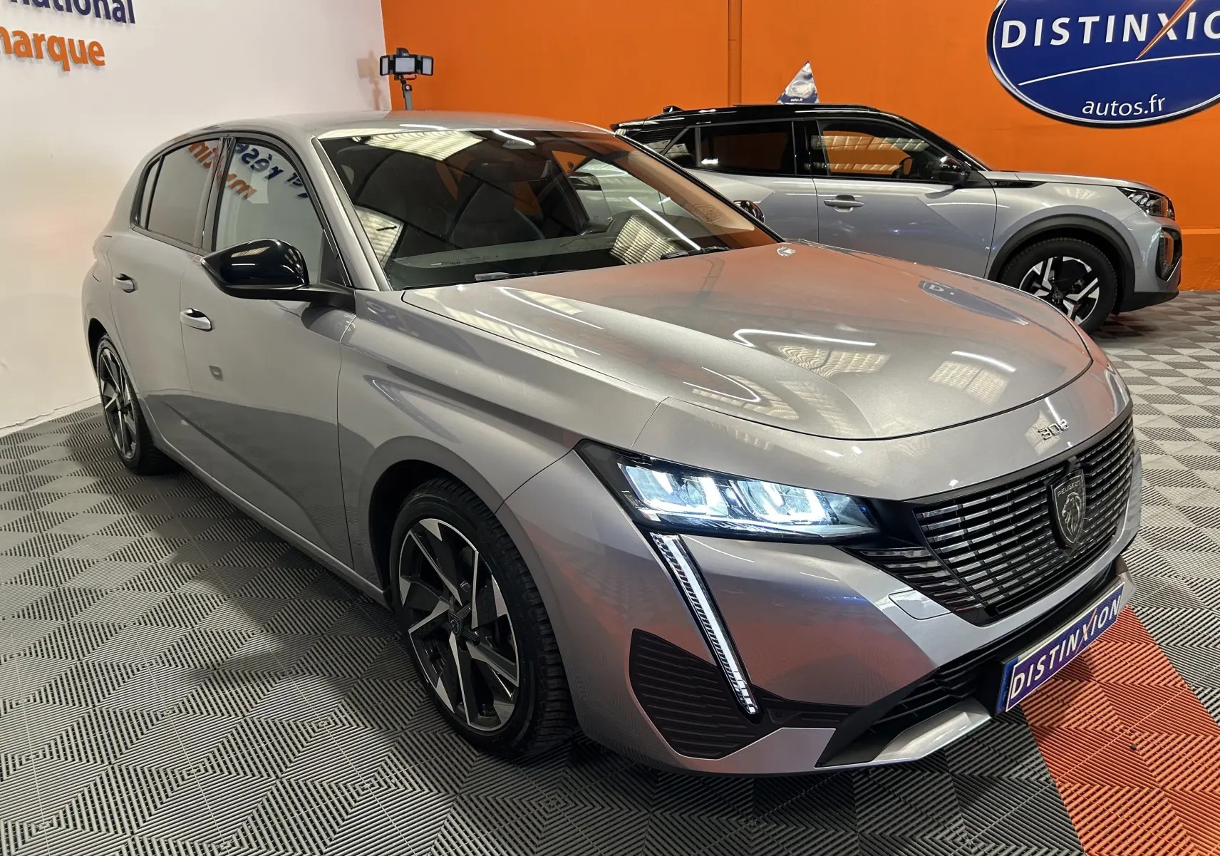 Peugeot 308 gris vue 3/4 avant droit, phares LED allumés, jantes alliage noires, en intérieur showroom.