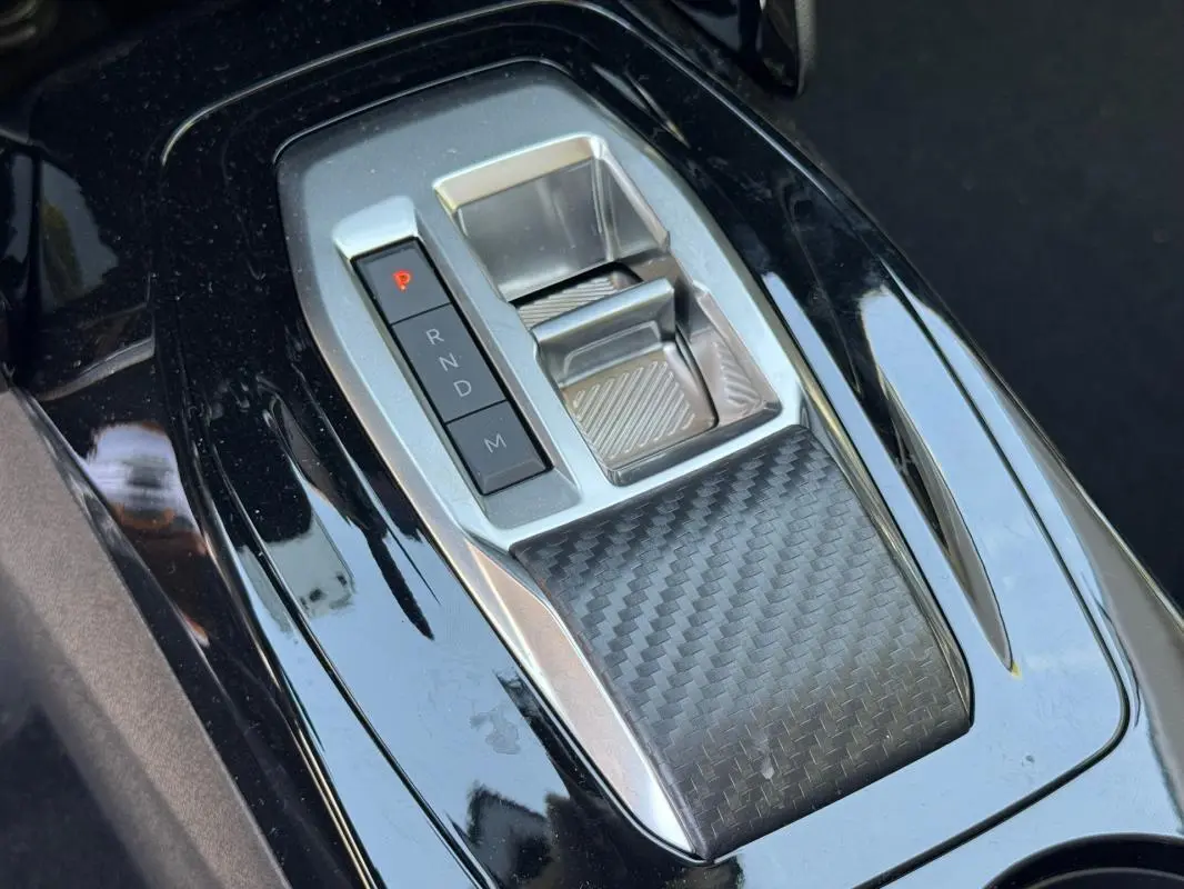 Gros plan sur la console centrale noire brillante de la Peugeot 208 Hybrid 110, avec sélecteur de boîte automatique et finition carbone.