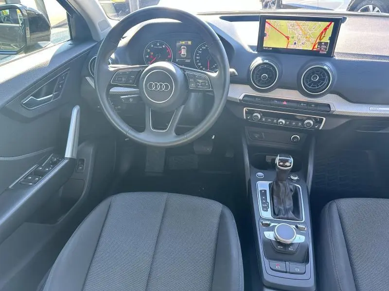 Vue intérieure avant de l'Audi Q2 blanc, montrant le volant cuir multifonction, l'écran GPS et la console centrale avec boîte auto.