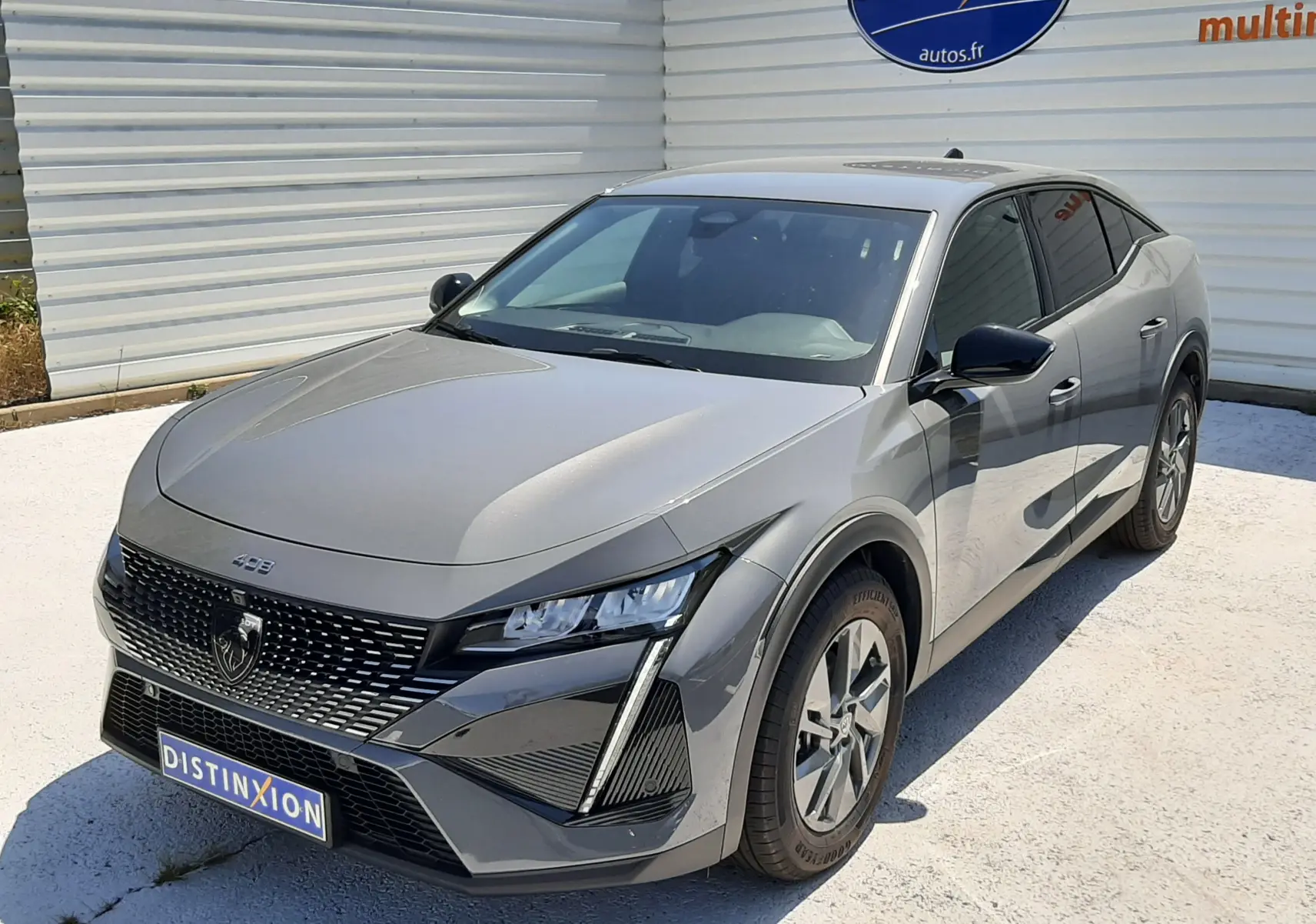 Peugeot 408 gris selenium vue 3/4 avant droit, avec calandre distinctive et phares LED anguleux, stationnée devant un mur blanc.