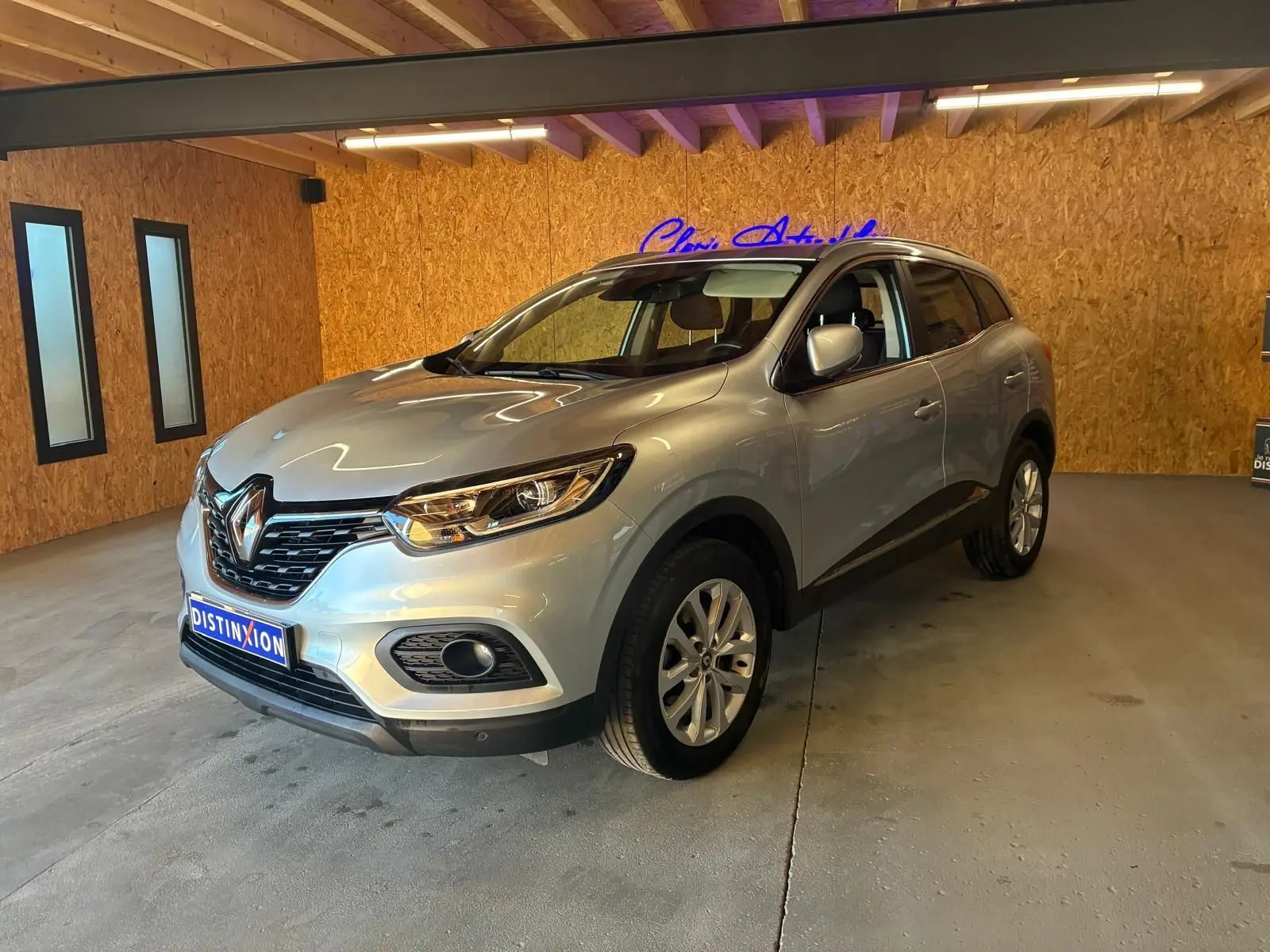 Renault Kadjar gris en 3/4 avant droit dans un garage avec éclairage intérieur et logo néon bleu en arrière-plan.