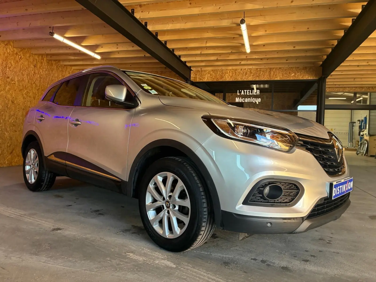 Renault Kadjar gris 3/4 avant droit, SUV compact avec jantes alliage et calandre noire, en intérieur d'atelier mécanique.