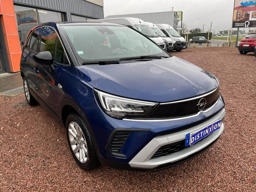 Vue 3/4 avant d'un Opel Crossland bleu clair avec toit noir, garé sur un sol en gravier devant un bâtiment.