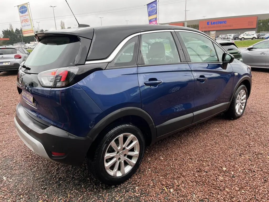 Vue 3/4 arrière droite d'un Opel Crossland bleu clair avec toit noir, garé sur un sol en gravier rouge.