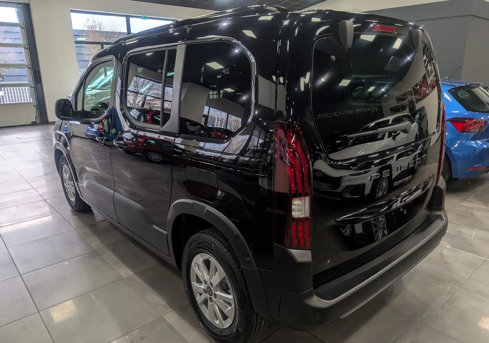 Vue 3/4 arrière droite d'un Peugeot Rifter noir 2025 en intérieur, avec feux arrière verticaux et jantes alliage.