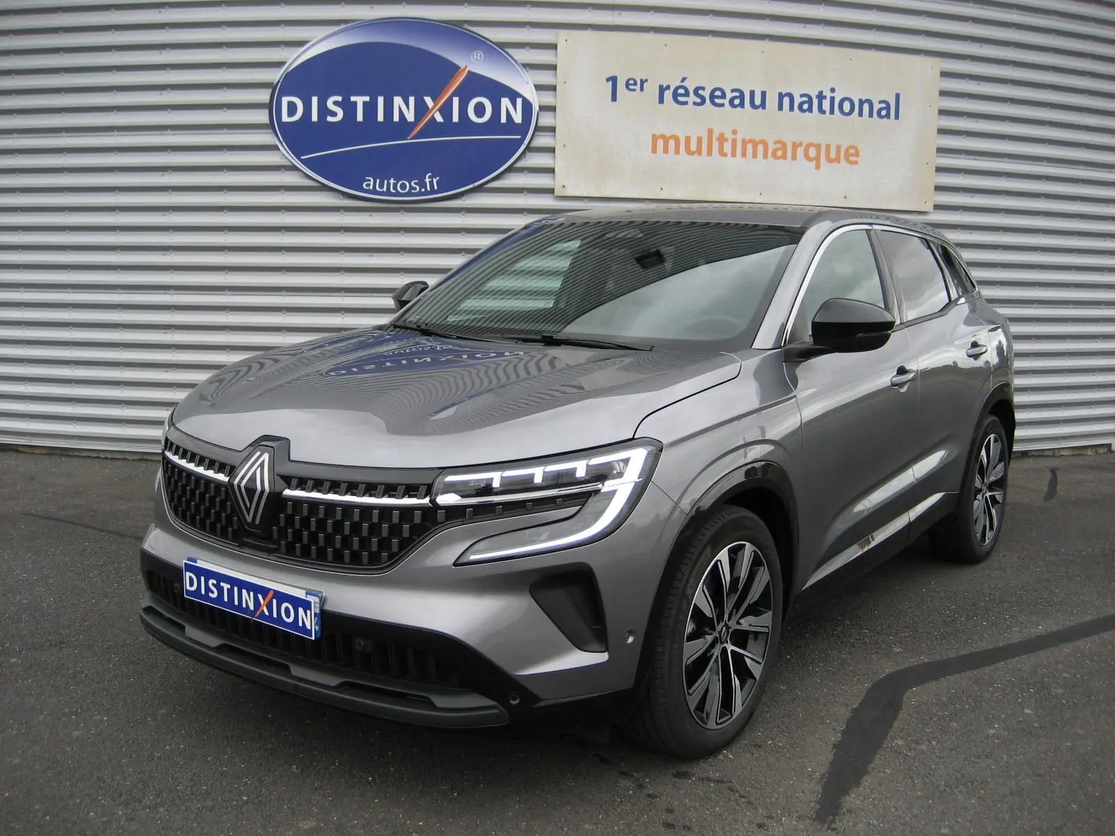 Renault Austral gris 3/4 avant droit, phares LED allumés, calandre noire et jantes bi-ton 19 pouces visibles.