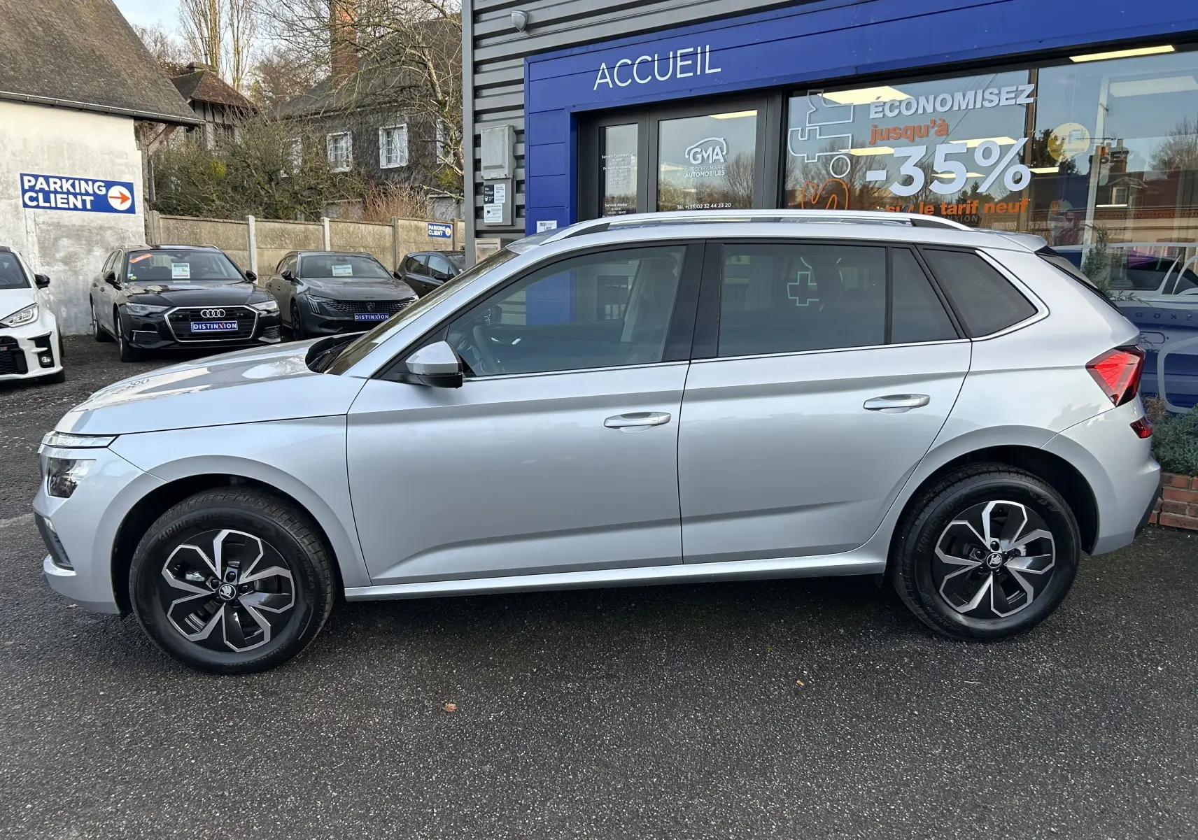 Vue latérale droite d’un Skoda Kamiq gris argent métallisé 2025, avec jantes alliage et vitres teintées, stationné devant un accueil.