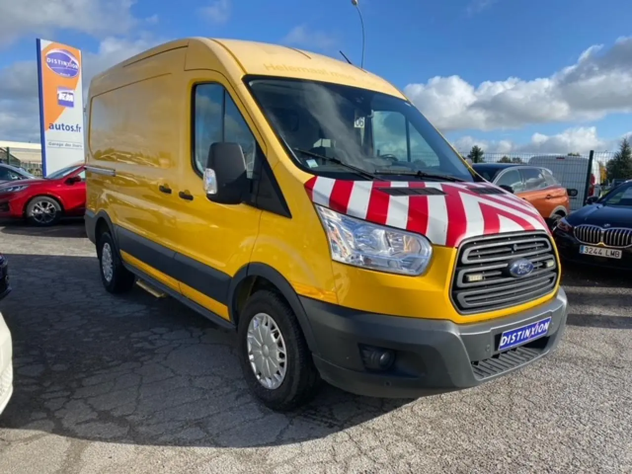 Ford Transit Fourgon orange vu en 3/4 avant droit avec capot rayé rouge et blanc sur un parking extérieur.