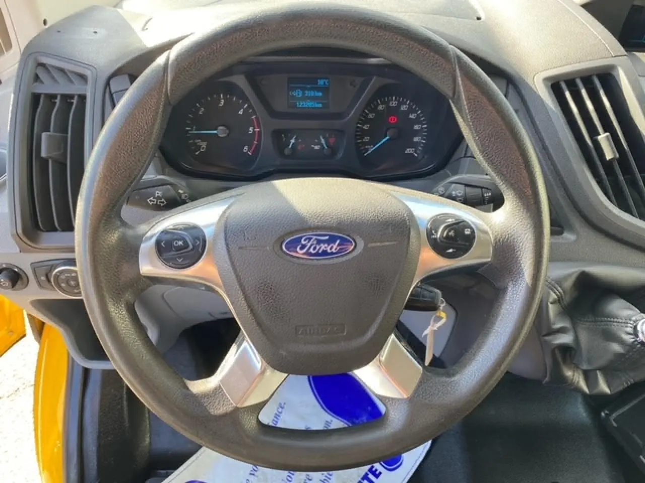 Vue rapprochée du volant Ford du Transit Fourgon orange 2015, avec commandes intégrées et tableau de bord visible.