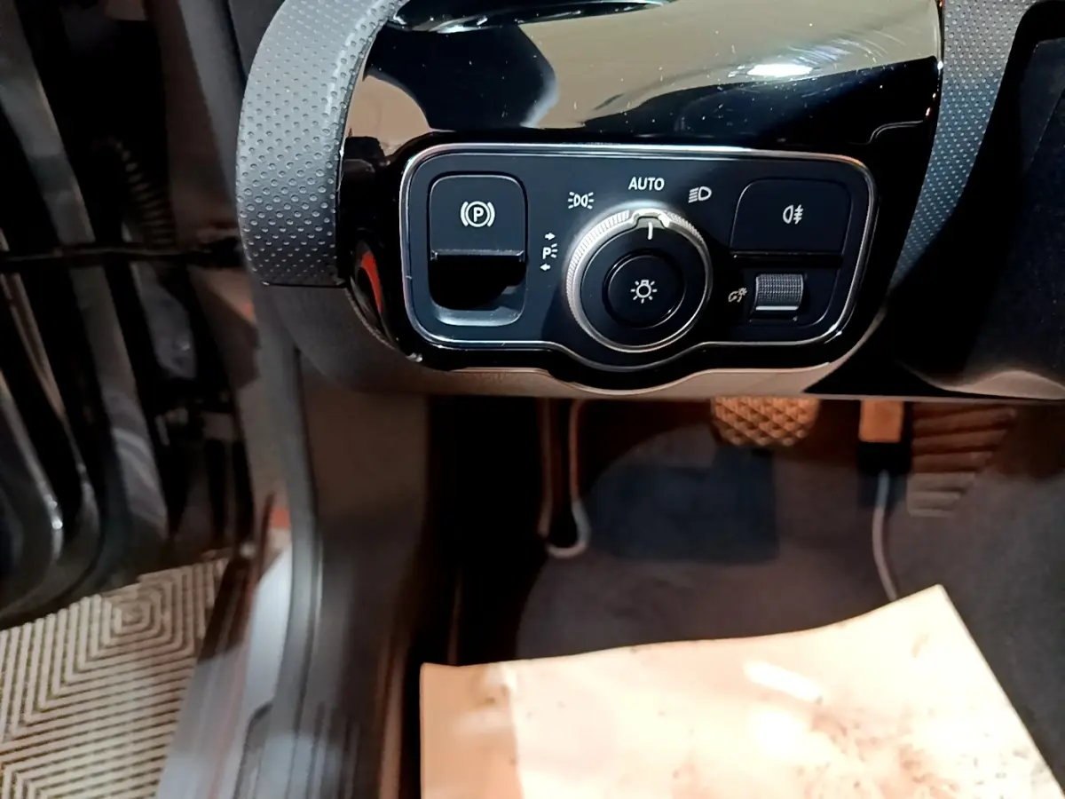 Gros plan sur les commandes d’éclairage et frein à main électrique côté gauche du tableau de bord noir brillant d’une Mercedes CLA 180 d gris clair.