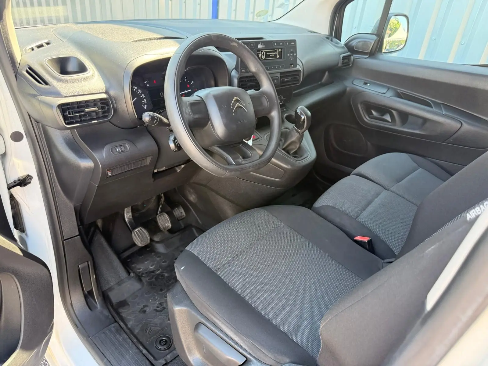 Vue intérieure côté conducteur du Citroën Berlingo blanc 2020, montrant le volant, tableau de bord et sièges tissu noirs.