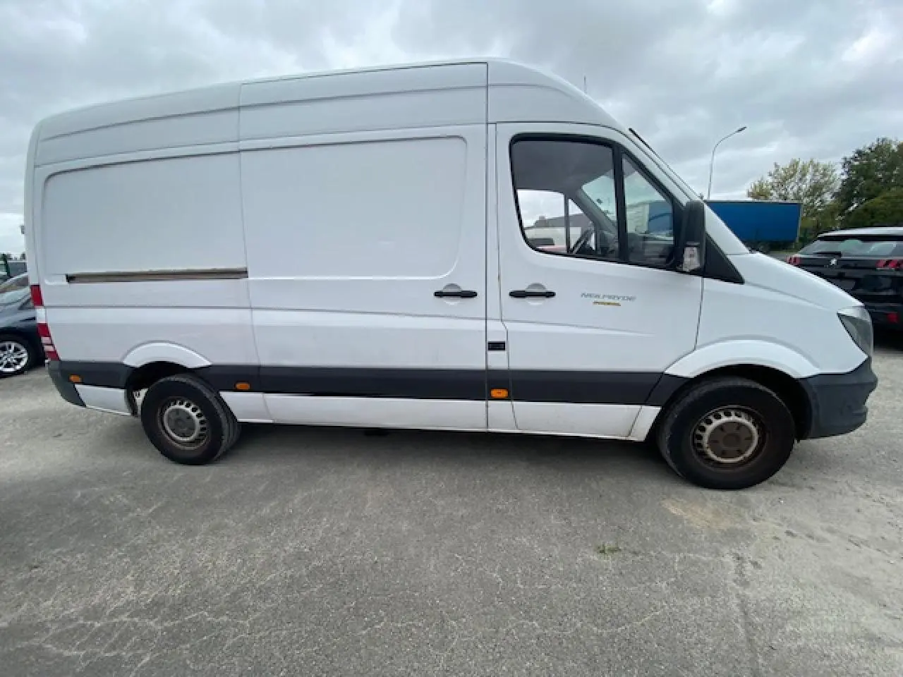 Profil côté droit d'un fourgon Mercedes Sprinter blanc de 2015 avec bande noire et jantes acier simples.