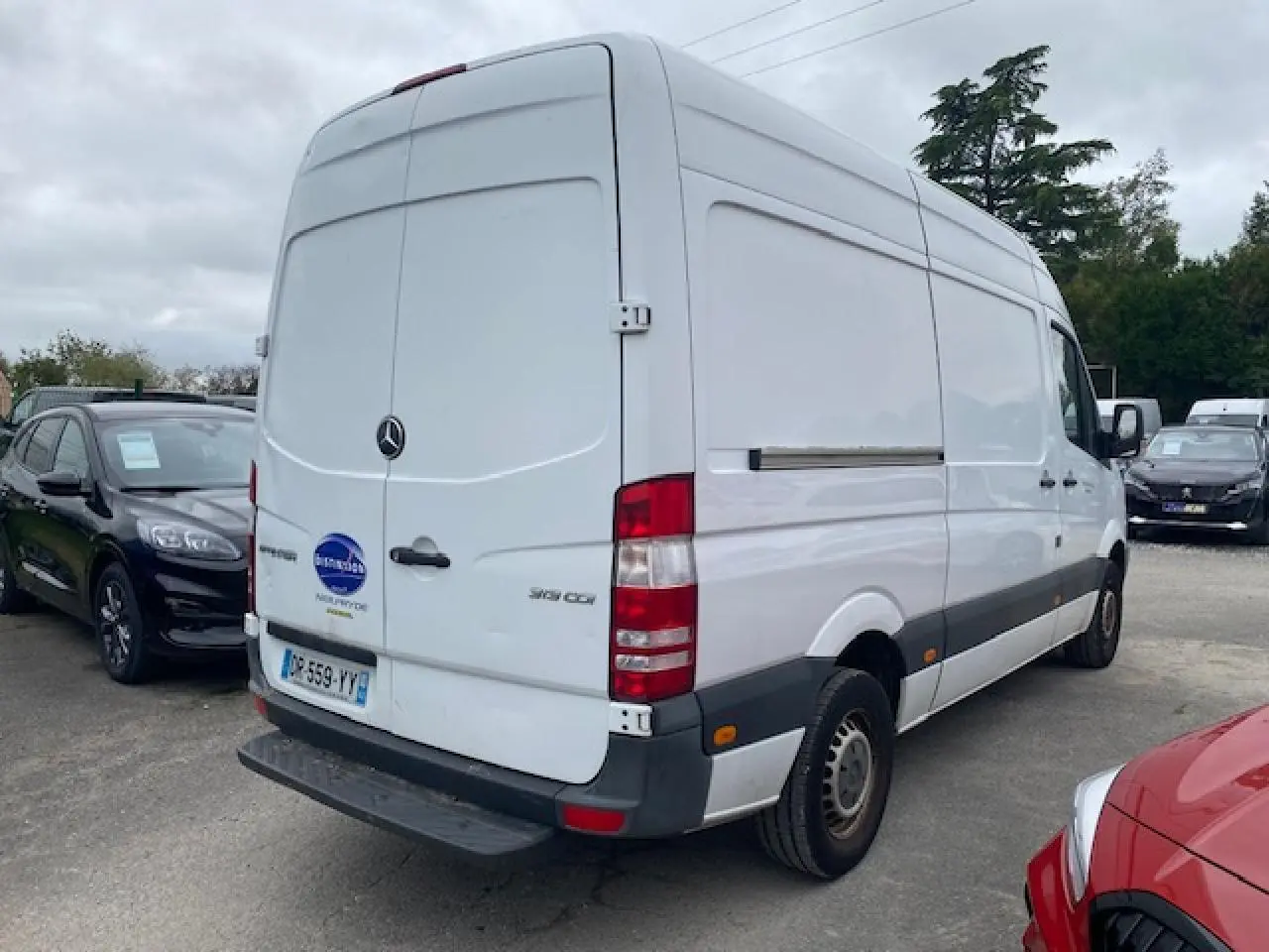 Vue 3/4 arrière droite d'un fourgon Mercedes Sprinter blanc de 2015 avec porte arrière fermée sur parking.