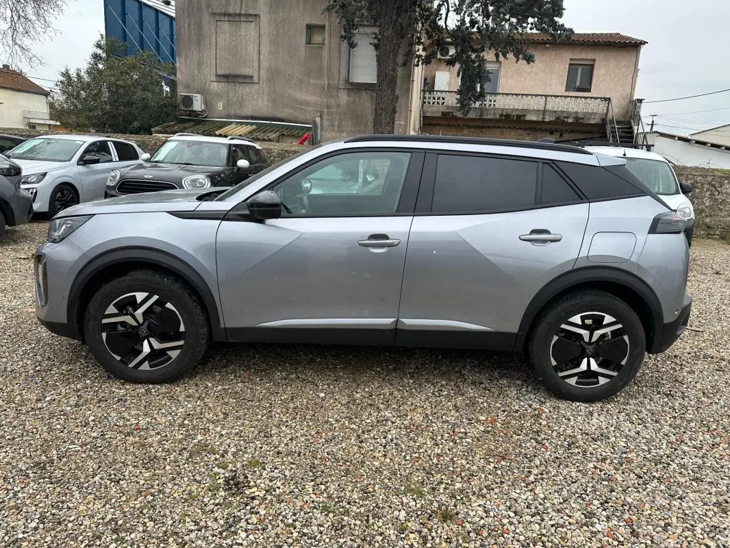 Vue de profil côté gauche d’un Peugeot 2008 Hybrid gris avec barres de toit et jantes alliage 17 pouces.