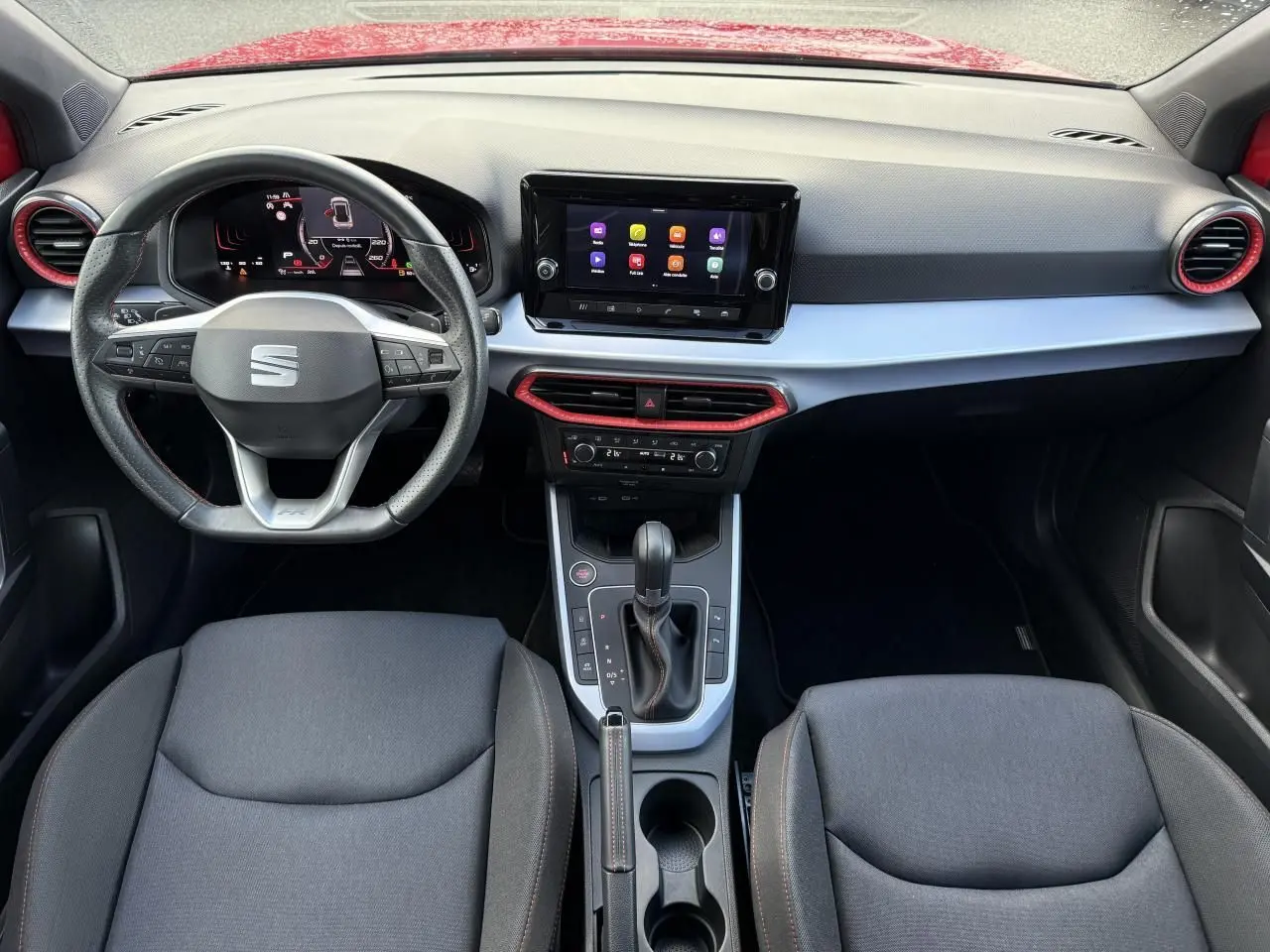 Vue intérieure avant du tableau de bord du SEAT Arona rouge, avec volant sport cuir perforé et écran tactile central.