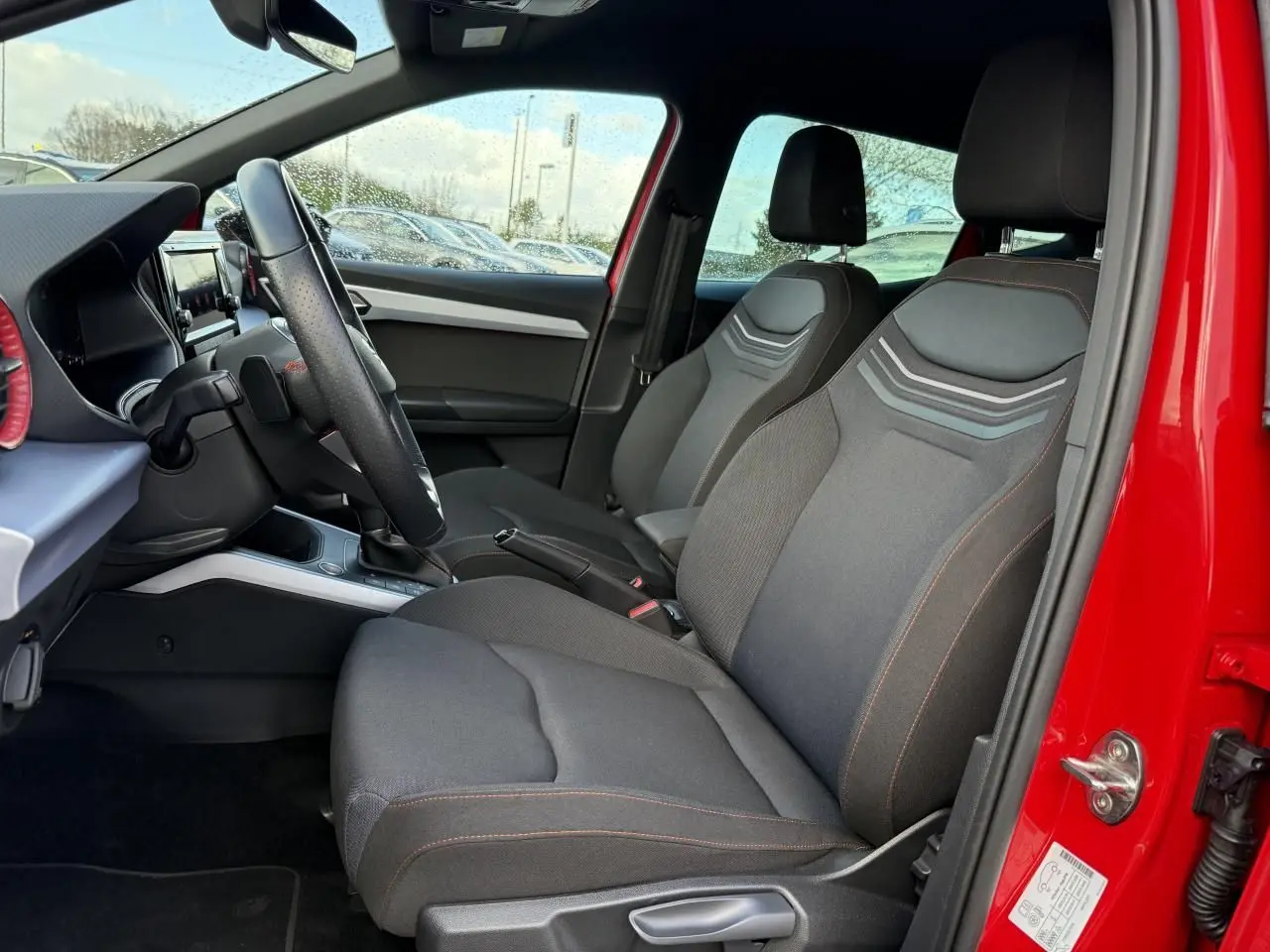 Vue intérieure côté conducteur du SEAT Arona rouge 2024, mettant en valeur les sièges avant noirs avec surpiqûres rouges et volant sport.