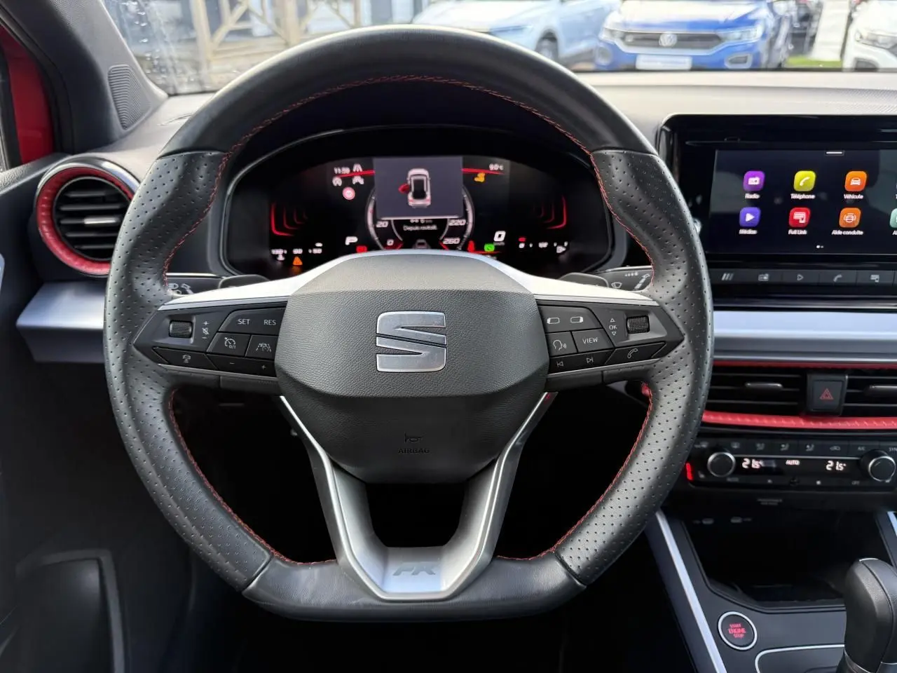 Vue rapprochée du volant sport en cuir perforé noir avec surpiqûres rouges et tableau de bord digital d'une SEAT Arona rouge 2024.