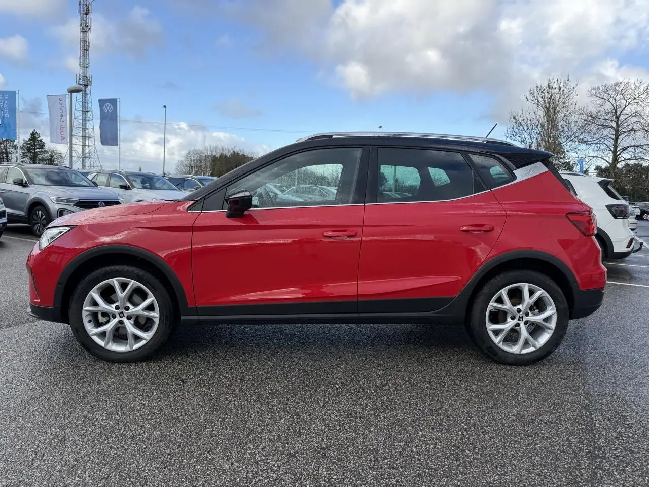 Vue de profil côté gauche d’un SEAT Arona rouge 2024 avec toit noir et jantes argentées sur parking.