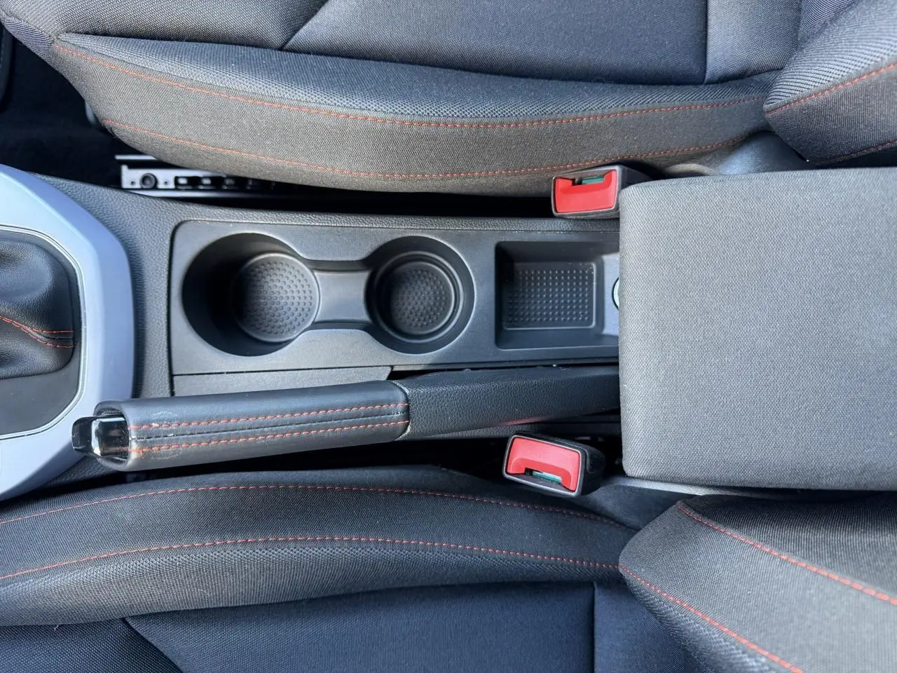 Vue plongeante sur la console centrale noire du SEAT Arona 2024 avec frein à main et porte-gobelets, surpiqûres rouges visibles.