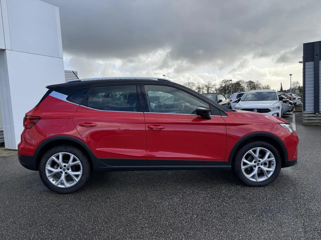 Profil droit d'un SEAT Arona rouge 2024 avec toit noir et jantes alliage argentées sur parking extérieur.