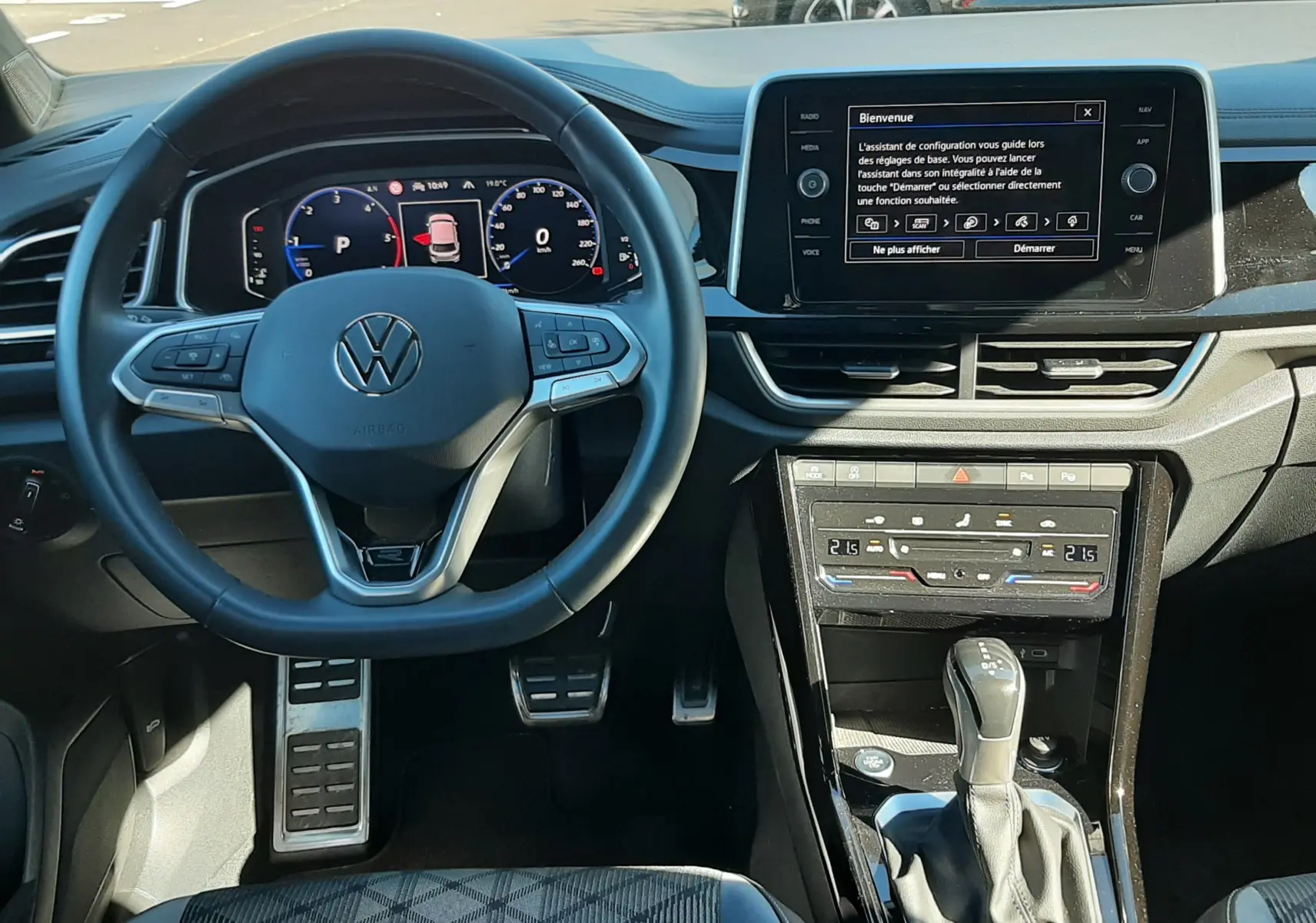 Vue intérieure du tableau de bord et volant du Volkswagen T-Roc gris Indium 2024, avec écran tactile et levier de boîte automatique.