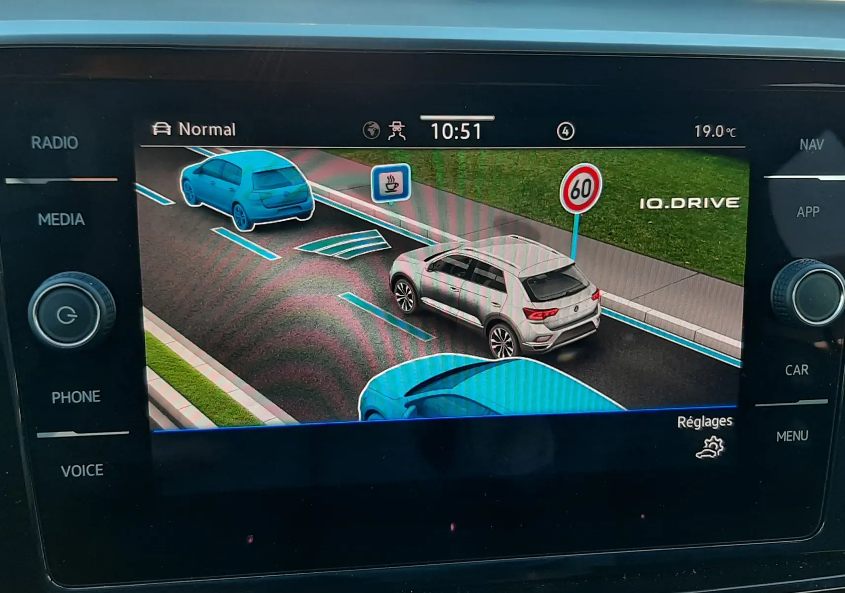 Affichage écran voiture montrant un Volkswagen T-Roc gris Indium en vue 3/4 arrière sur une route avec limitation à 60 km/h.