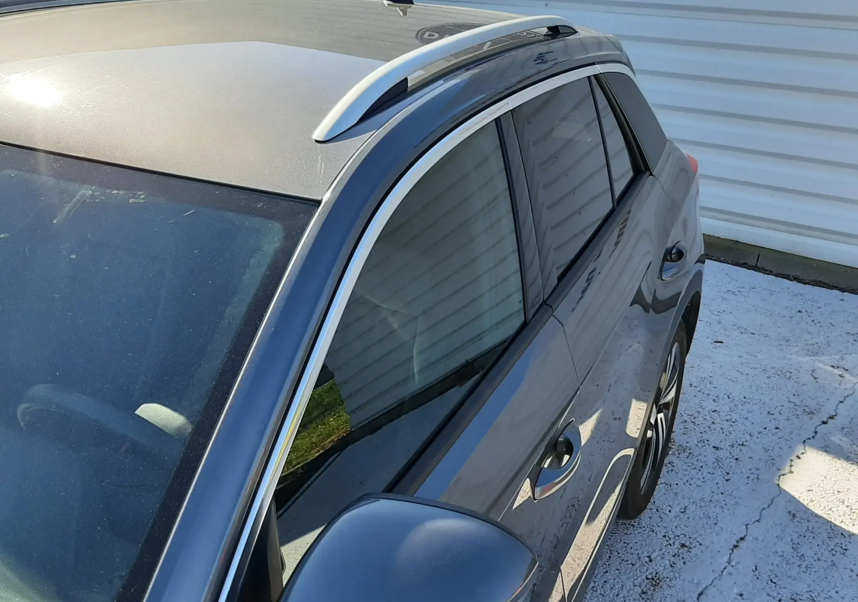 Vue en plongée du côté gauche d’un Volkswagen T-Roc gris Indium avec barres de toit et rétroviseur assorti.