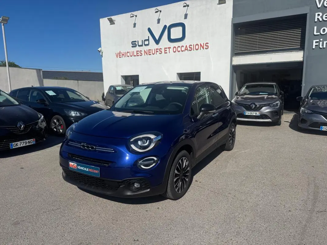 Fiat 500X bleu en 3/4 avant droit, avec jantes classiques et phares ronds distinctifs, stationnée devant un concessionnaire.