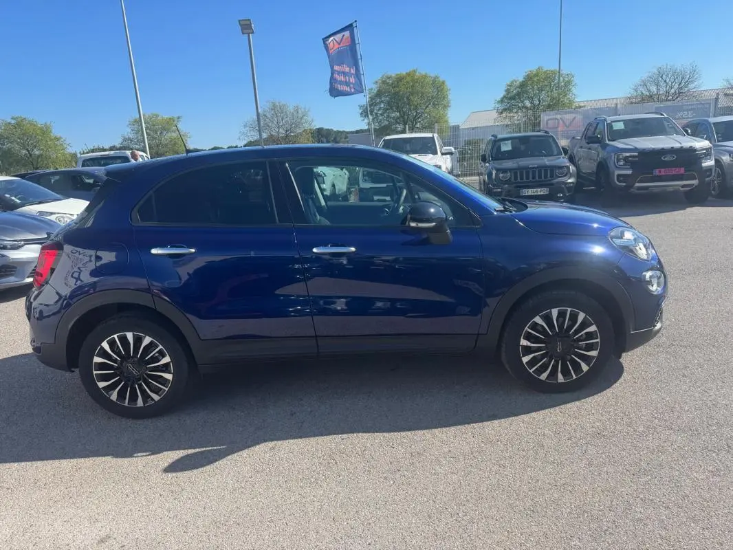 Profil côté gauche d'un FIAT 500X bleu MY23 avec jantes classiques 16 pouces, garé en extérieur par temps clair.