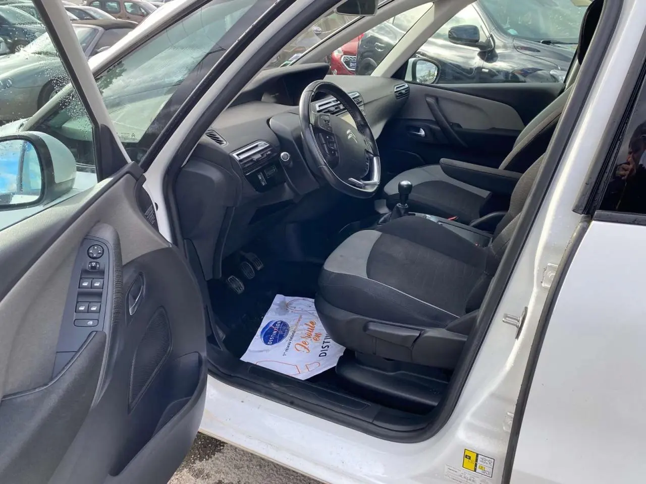 Intérieur côté conducteur d'un Citroën Grand C4 Picasso blanc, montrant le volant cuir, levier de vitesse manuel et sièges tissu gris.
