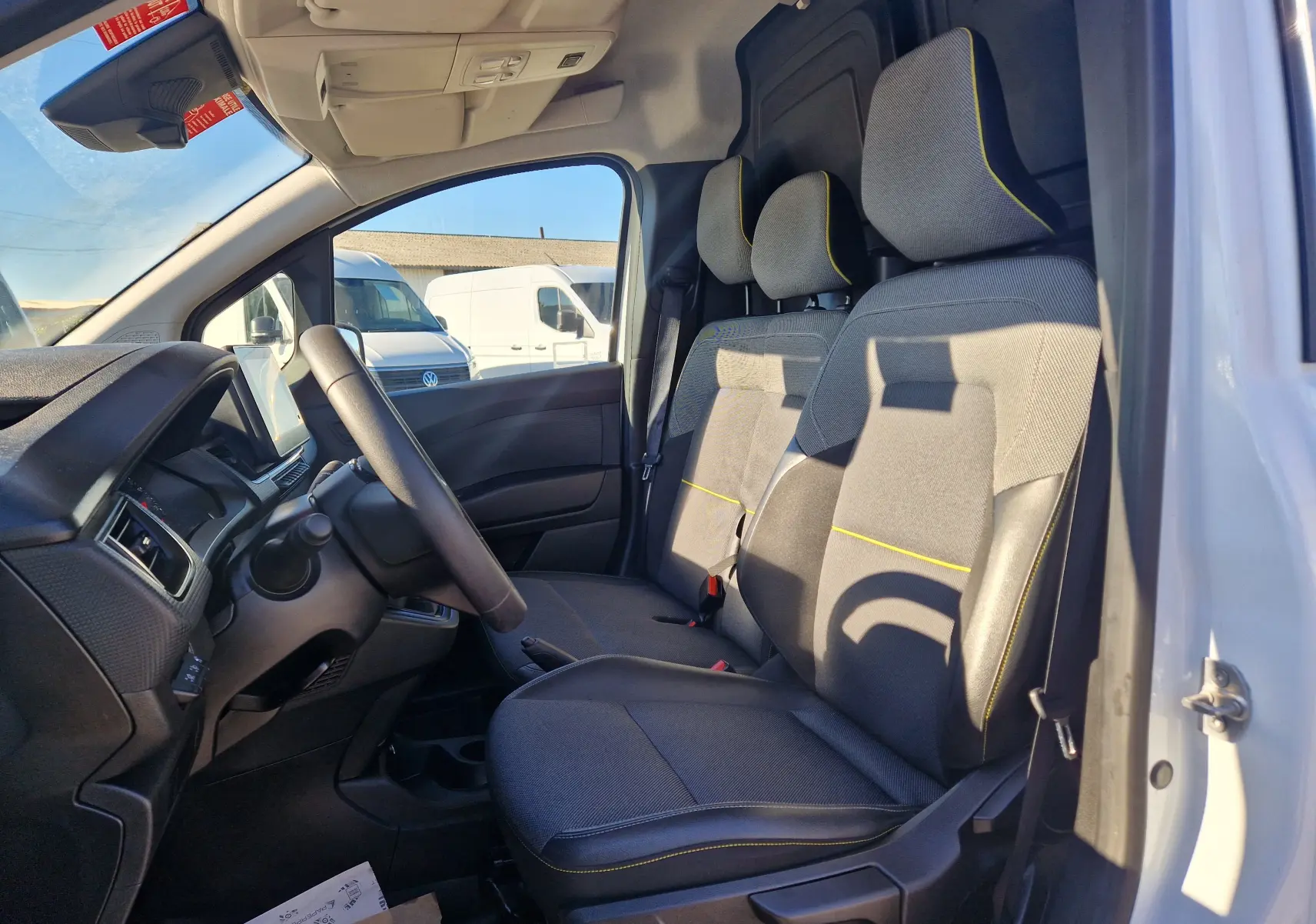 Vue intérieure côté conducteur du Renault Kangoo blanc 2024, montrant les sièges gris avec surpiqûres jaunes et le tableau de bord.