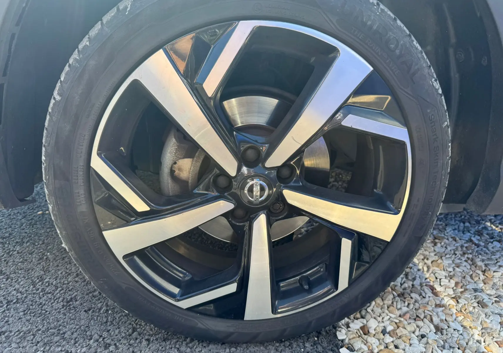 Gros plan sur la jante alu noire et argentée du Nissan Qashqai II PH2 2017 avec pneu Uniroyal visible sur sol gravillonné.