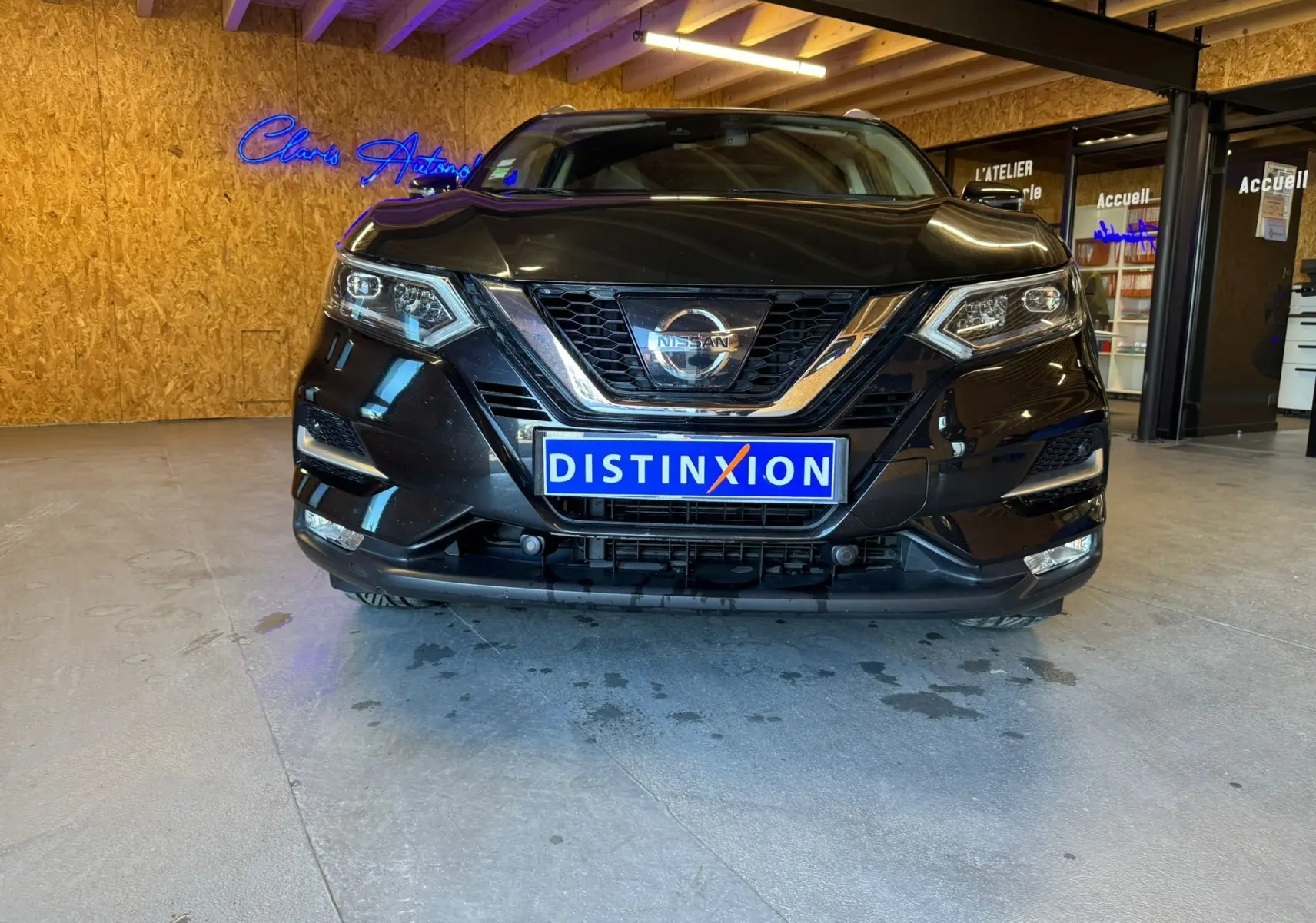 Vue avant d'un Nissan Qashqai II PH2 noir avec calandre chromée et plaque Distinxion en intérieur d'atelier.