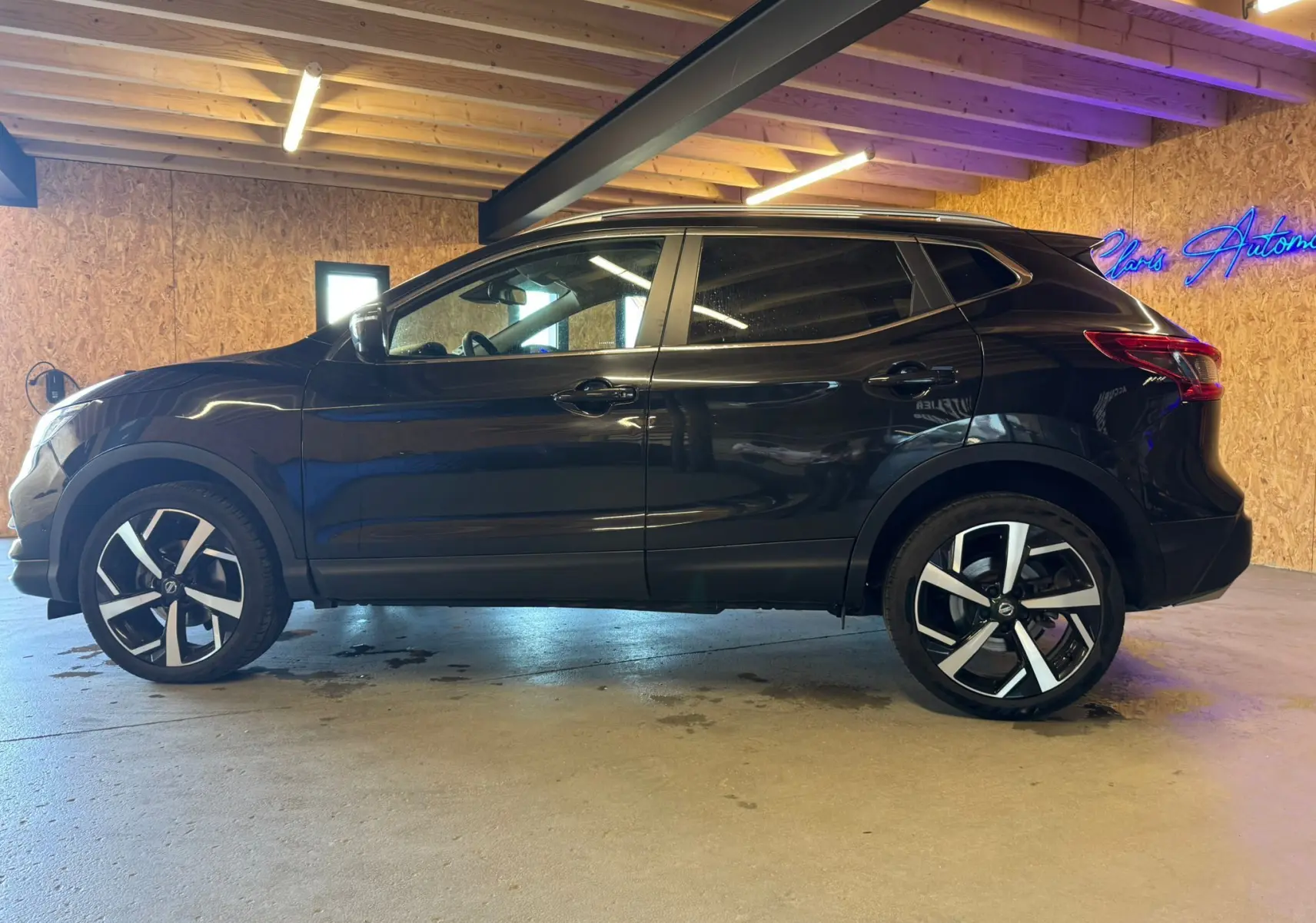 Vue de profil côté gauche d'un Nissan Qashqai noir 2017 avec jantes alu et barres de toit en intérieur garage.