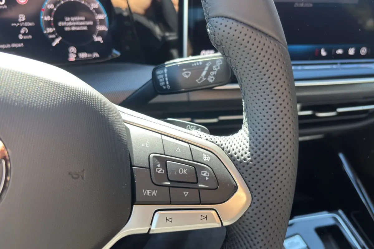 Gros plan sur le volant cuir perforé côté gauche d’une Volkswagen Golf blanche avec commandes multifonctions et tableau de bord numérique.