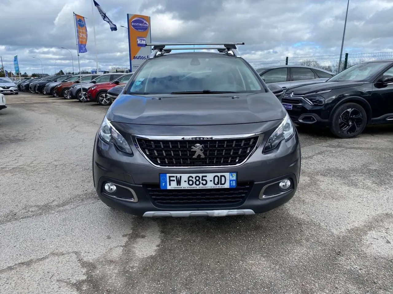 Vue frontale d'un Peugeot 2008 gris platinium avec barres de toit aluminium sur un parking extérieur.