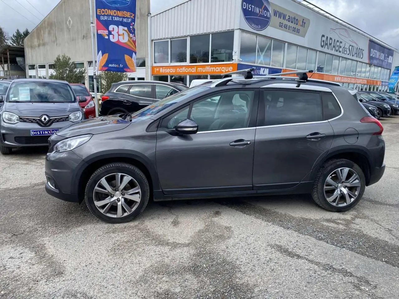 Profil côté gauche du Peugeot 2008 gris platinium 2019 avec barres de toit aluminium sur parking extérieur.