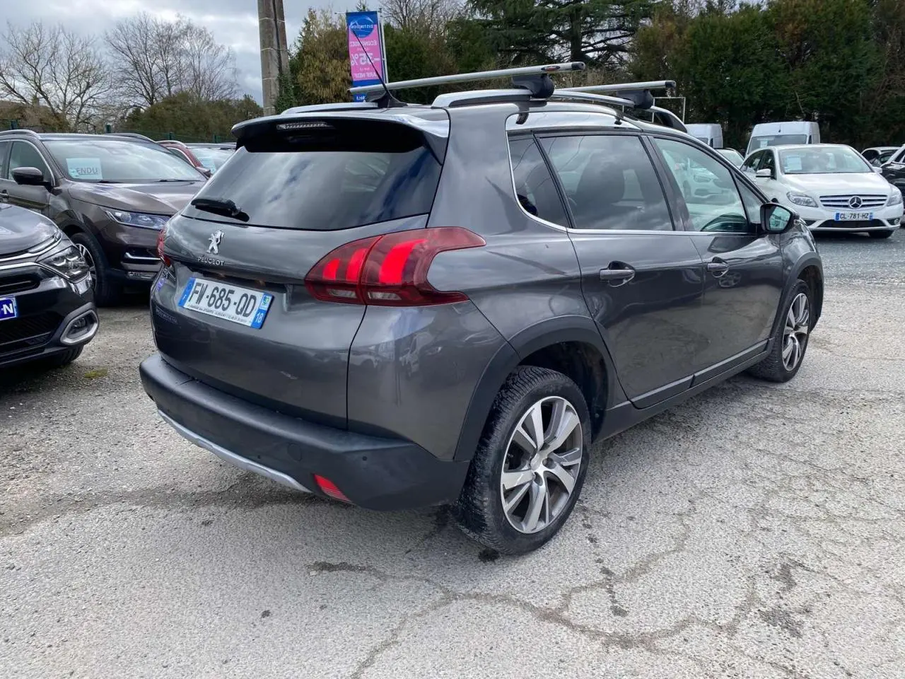 Peugeot 2008 gris platinium vue 3/4 arrière droit avec barres de toit aluminium sur parking.
