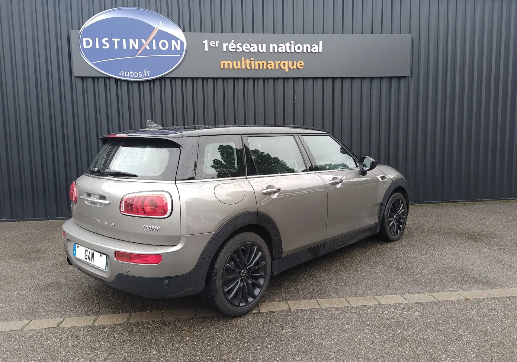 Vue 3/4 arrière droite d'une MINI Clubman F54 Silver Metal avec toit noir et jantes noires, stationnée devant un mur gris.