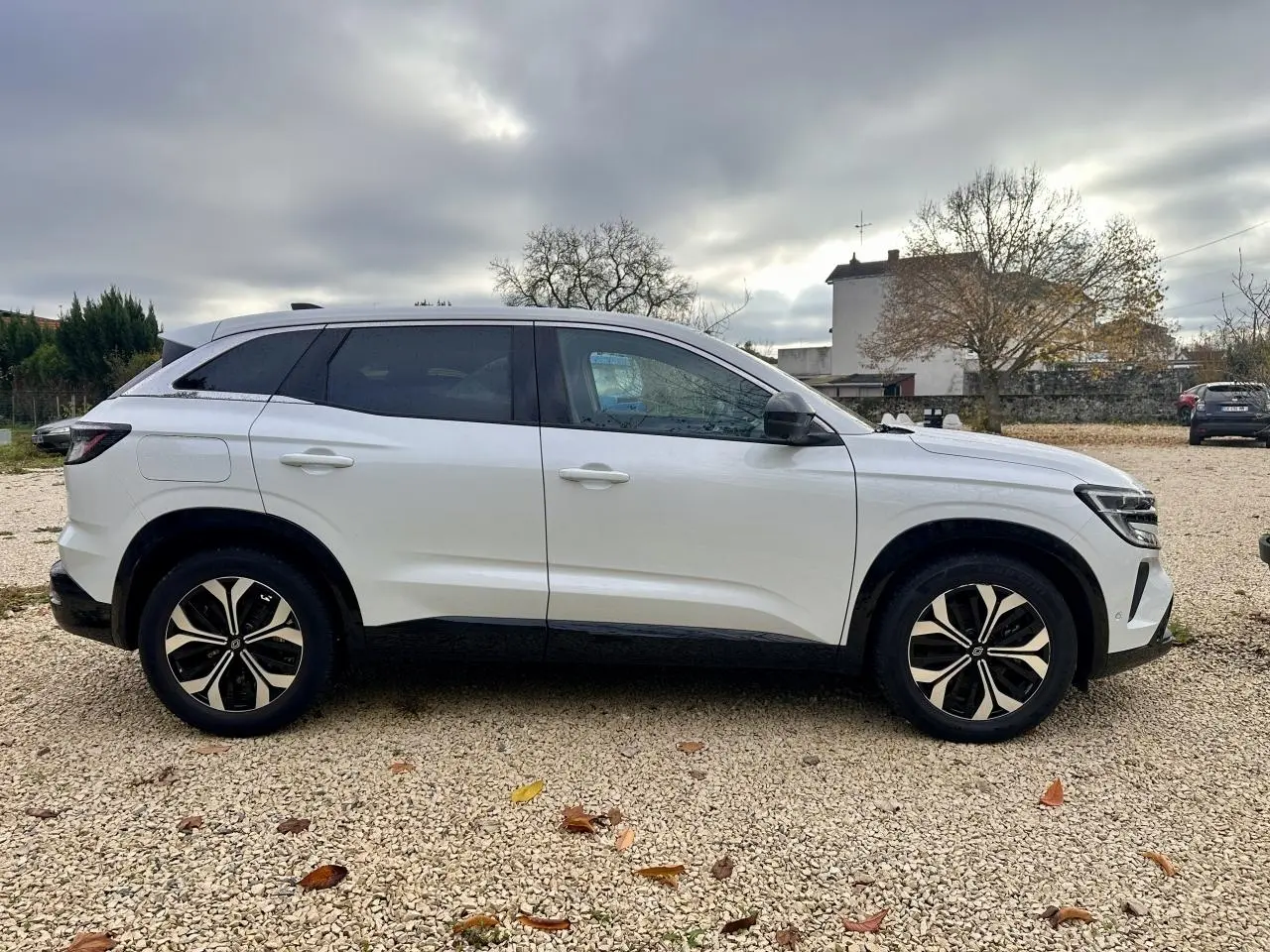Profil côté gauche d'un Renault Austral blanc 2024 avec jantes noires et toit noir sur fond de ciel nuageux.