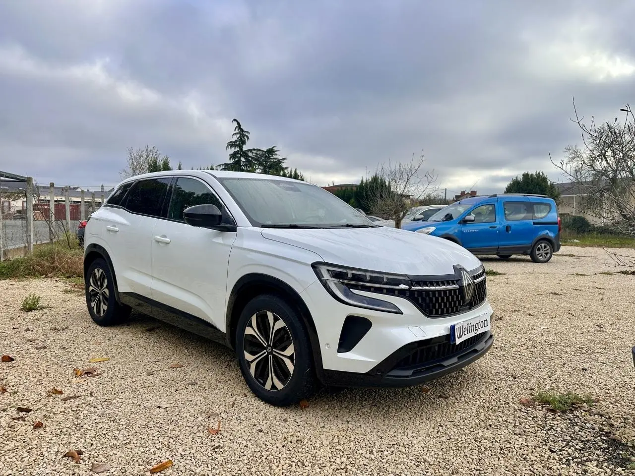 Renault Austral blanc en 3/4 avant droit, avec calandre noire et jantes modernes sur un sol gravillonné.