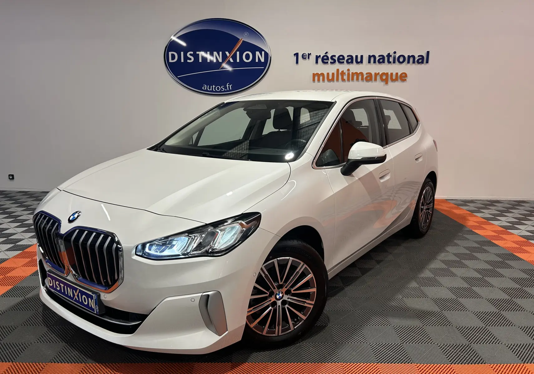 BMW Série 2 Active Tourer blanc vue 3/4 avant droit en intérieur avec calandre imposante et phares allumés.