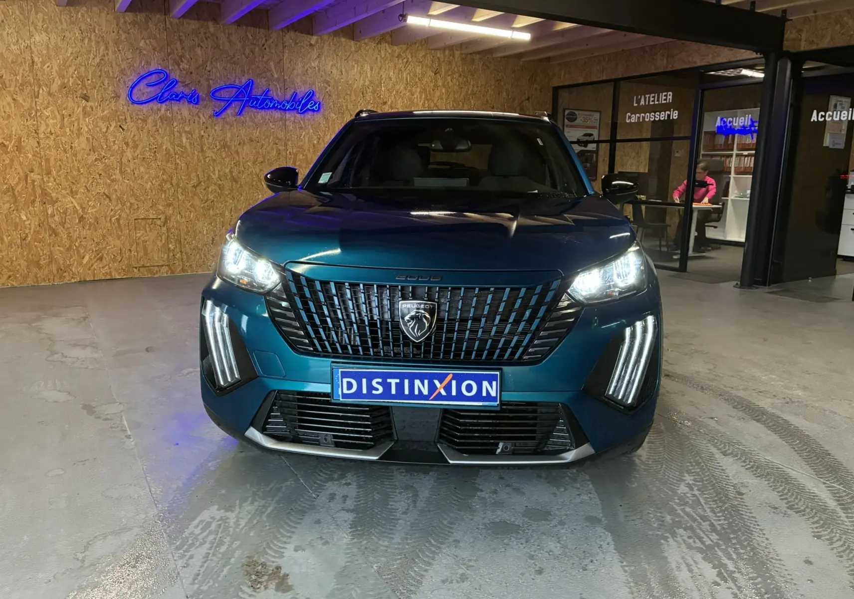 Vue de face d'un Peugeot 2008 bleu 2025 avec phares allumés et calandre distinctive en intérieur d'atelier.
