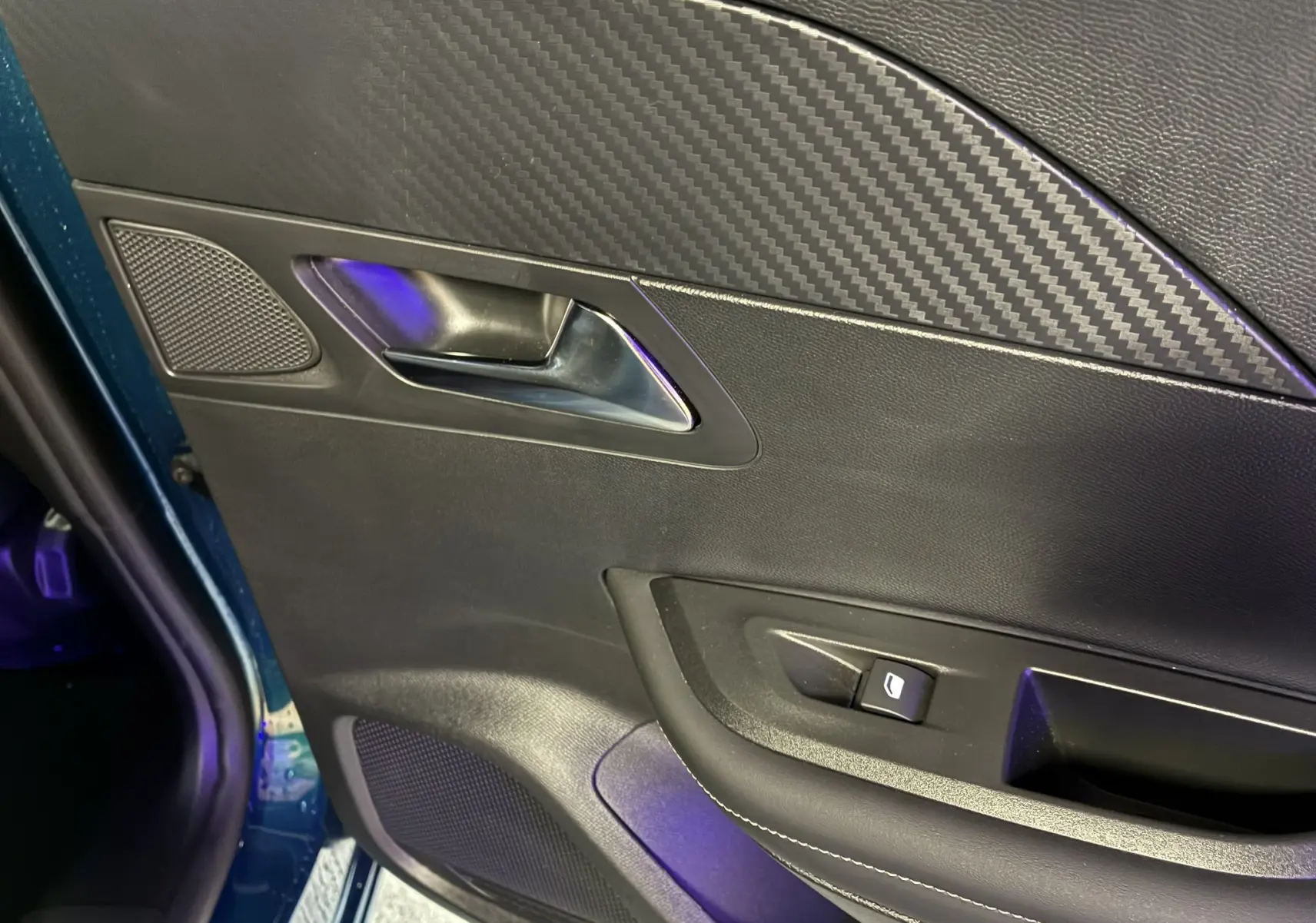 Détail de la porte intérieure côté gauche d’un Peugeot 2008 bleu 2025, avec poignée métal et insert effet carbone.