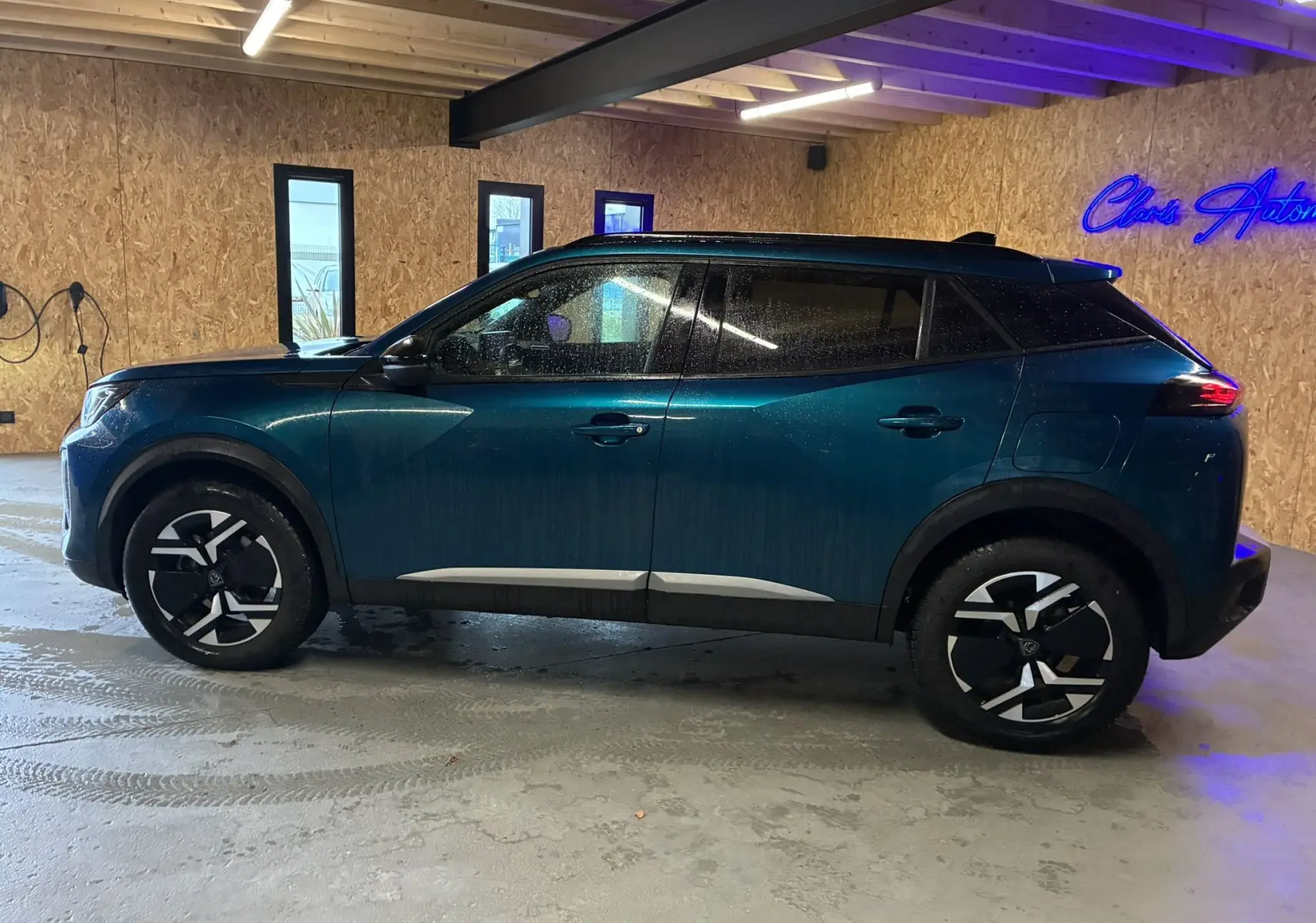 Profil latéral droit d'un Peugeot 2008 bleu hybride 2025, avec jantes noires et toit noir contrastant en intérieur d'atelier.